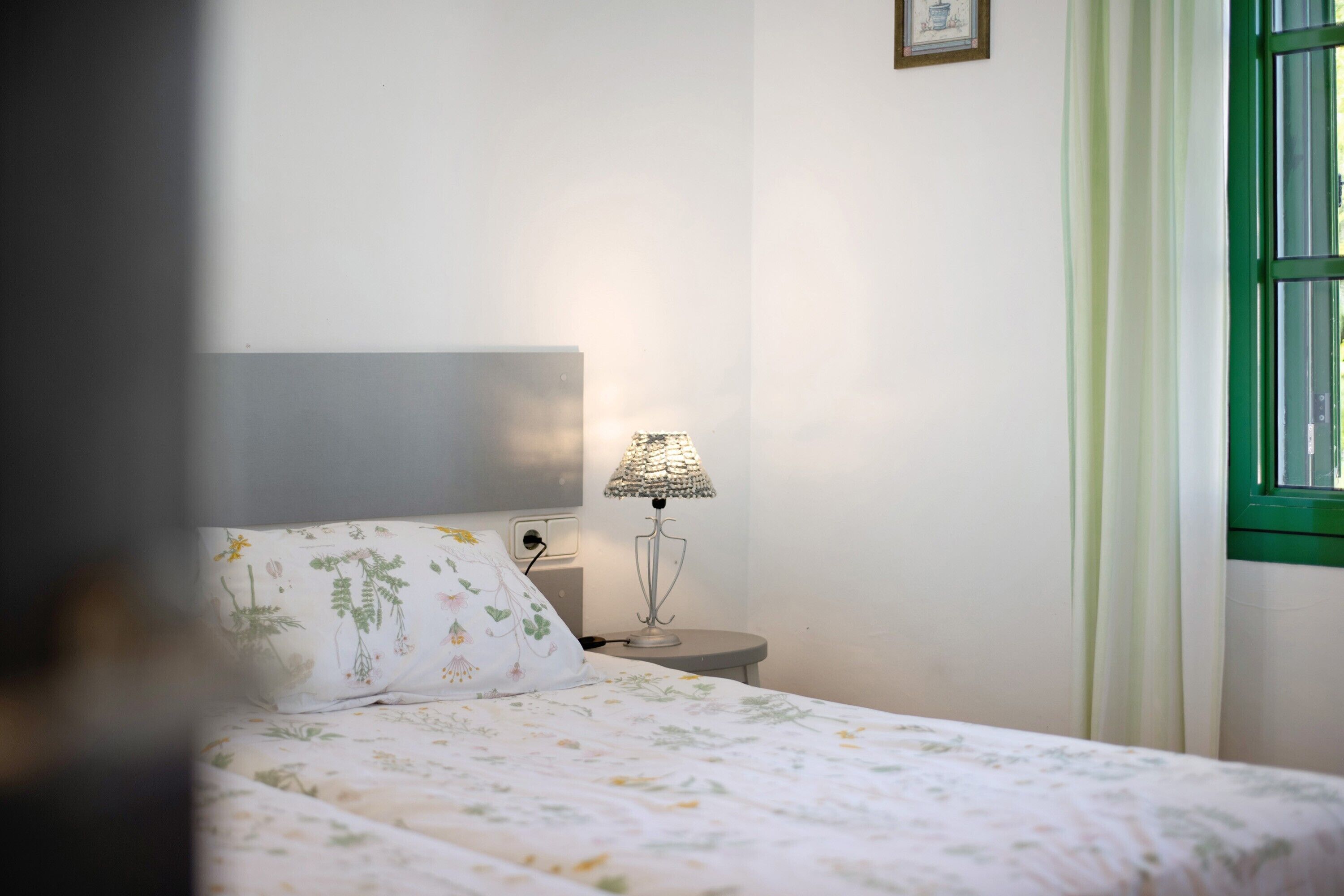 A bedroom with a bed, a nightstand, a lamp, a window with curtains, and a wall-mounted picture.