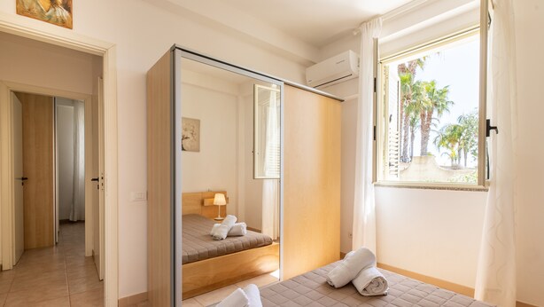 Une chambre d’hôtel avec un lit, un miroir, une fenêtre avec des rideaux, une porte et un couloir.
