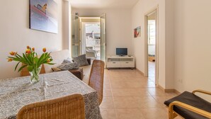 Un coin repas comprenant une table, des chaises, un vase de fleurs, un téléviseur posé sur un meuble, et un couloir menant à une chambre.