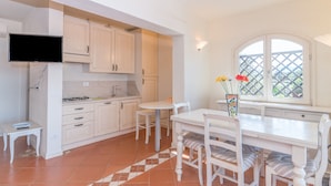Une cuisine avec des meubles blancs, une table à manger avec des chaises, un téléviseur fixé au-dessus du comptoir et une fenêtre ornée d’un treillis.