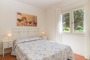 A bedroom with a bed, two bedside tables, a nightstand, a painting, and a window with curtains.