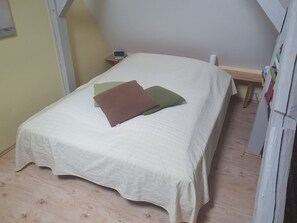 Una cama individual con cubrecama blanco acolchado, una mesita de noche de madera con un teléfono móvil y suelo de madera.