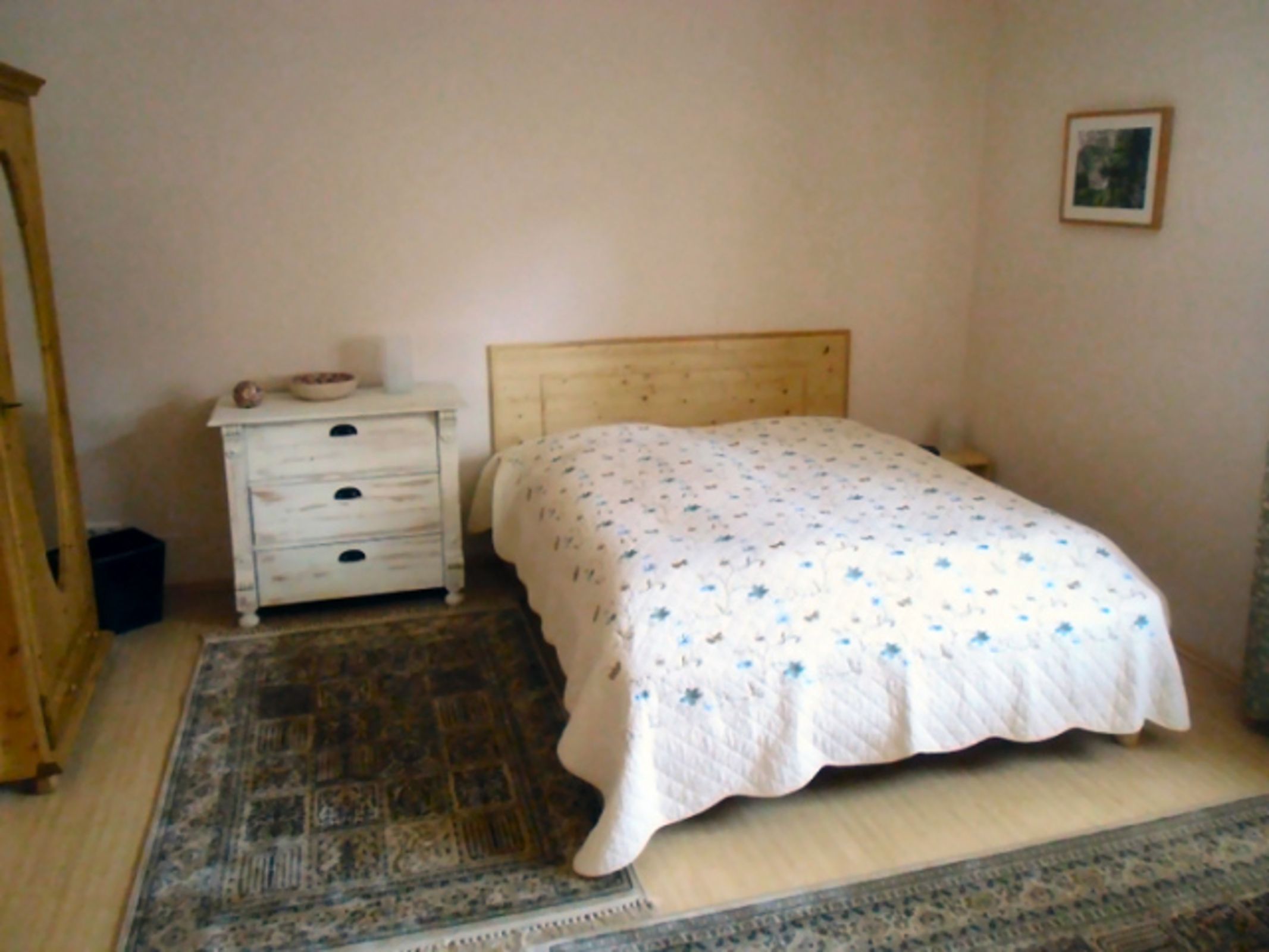 Una camera da letto con un letto in legno, un comodino bianco con cassetti, un tappeto decorato e un quadro appeso al muro.