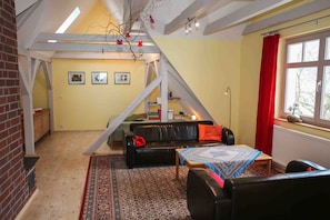 Ein Loft-Wohnraum mit schräg versiegelter Decke, einer schwarzen Ledersofa, einem gemusterten Teppich und einem kleinen Tisch mit blau-weißer Tischdecke.