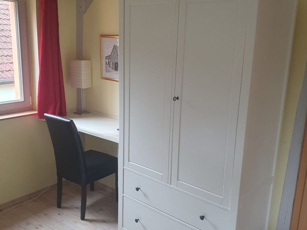 Ein Zimmer mit Schreibtisch, Stuhl, Kleiderschrank, Fenster mit roten Vorhängen und einer Lampe.