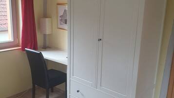 Ein Zimmer mit Schreibtisch, Stuhl, Kleiderschrank, Fenster mit roten Vorhängen und einer Lampe.