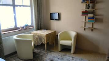 Ein Zimmer mit Fenster, Fernseher, kleinem Tisch, Stuhl und Bücherregal.