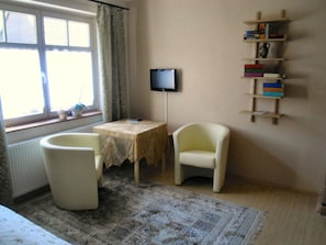 A room with a window, a television, a small table, a chair, and a bookshelf.