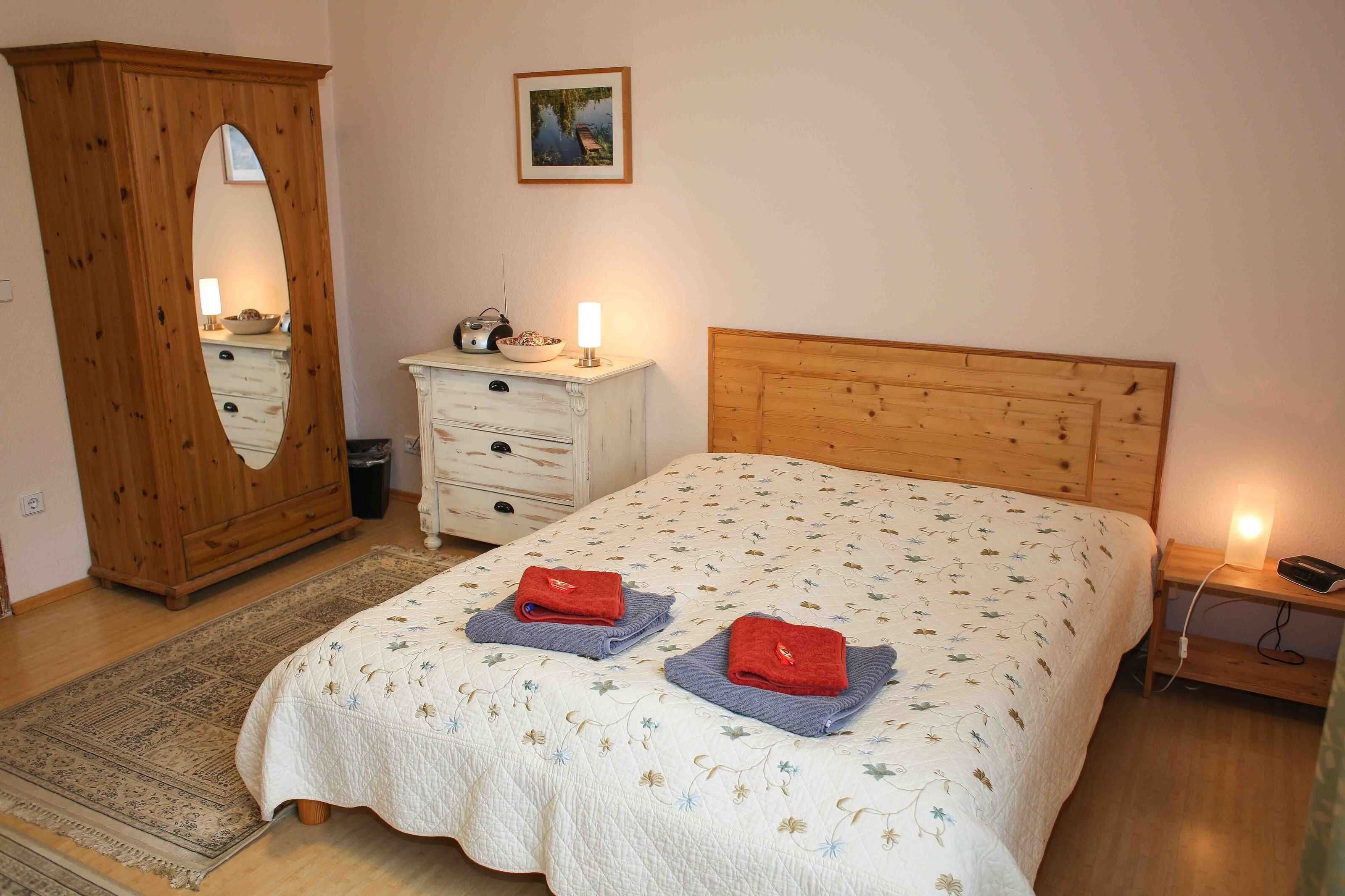 Ein Schlafzimmer mit einem hölzernen Kopfende, einem Bett mit floralem Bettzeug, zwei Handtüchern auf dem Bett, einem Holzschrank, einem Nachttisch mit einer Lampe und einem Bild an der Wand.