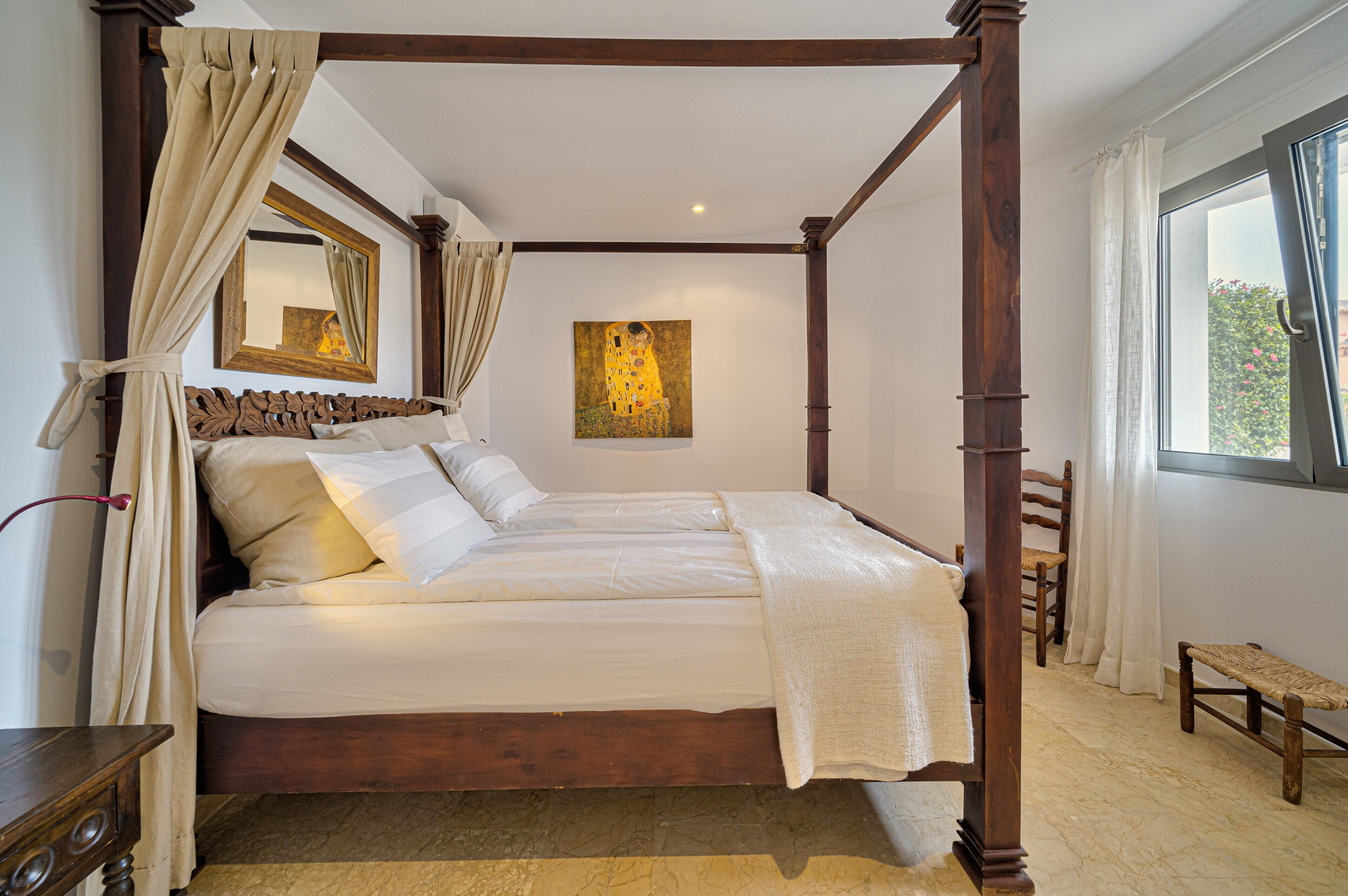 Una cama con dosel, un cabecero de madera, una mesita de noche, una silla y una ventana con cortinas.