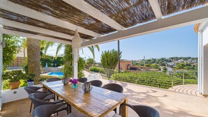 Un porxo cobert amb una taula de fusta i cadires de vimet, amb vistes a una piscina i a un paisatge idíl·lic d'una vila.