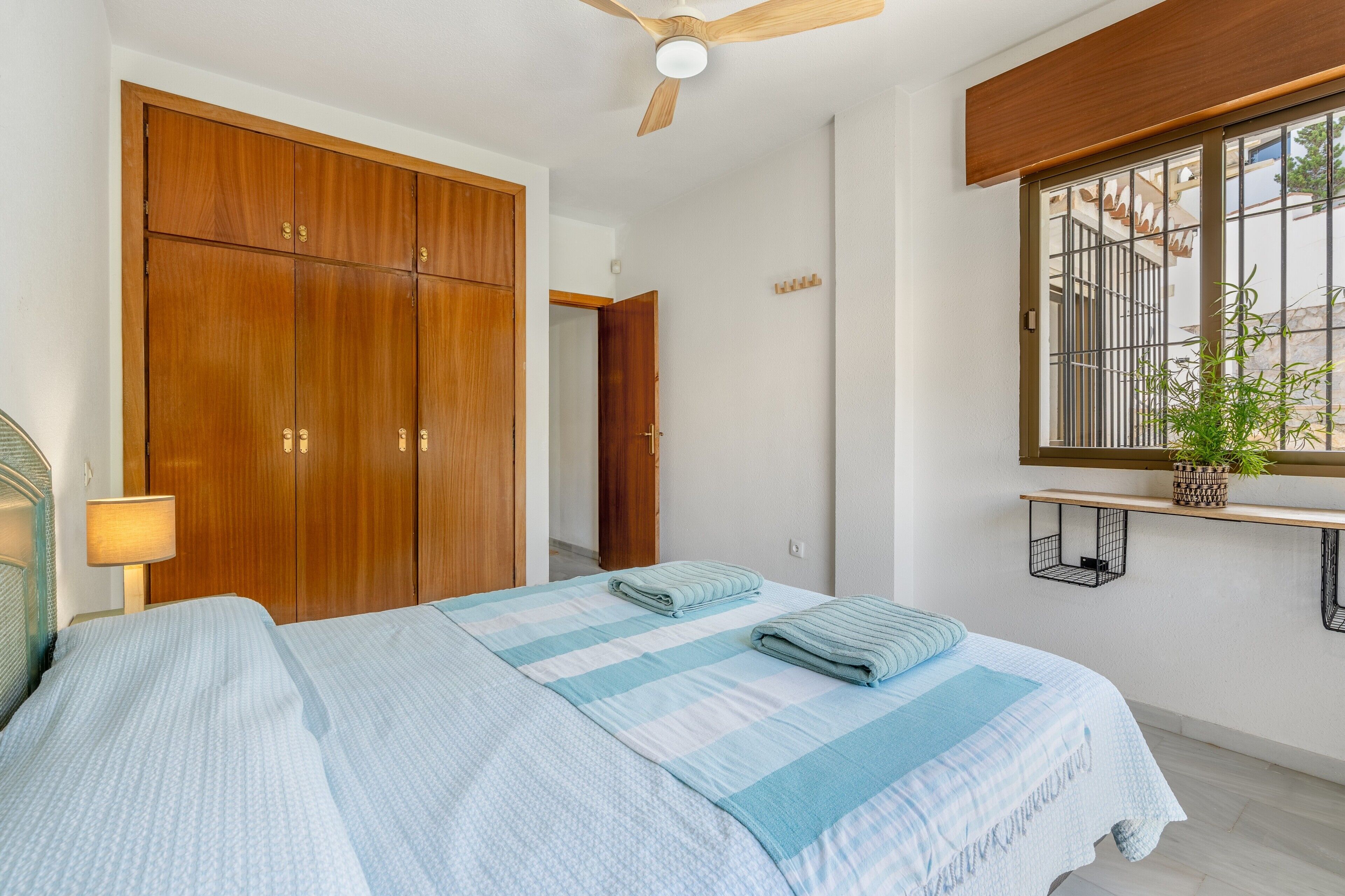 Un dormitorio con cama, armario de madera, ventilador de techo, ventana con vistas y planta en la mesita de noche.