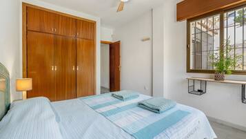 Un dormitorio con cama, armario de madera, ventilador de techo, ventana con vistas y planta en la mesita de noche.
