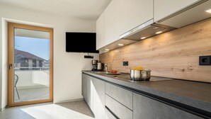 A modern kitchen with a wooden backsplash, a flat-screen TV mounted above the counter, and a sliding glass door leading to a balcony. - Apartment "Sunnig - Sonnenaufgang" with Mountain View, Balcony & Wi-Fi (Völlan)