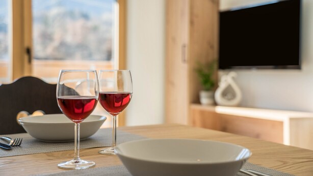 A dining table with two wine glasses and a bowl, a wooden cabinet, and a TV mounted on the wall. - Apartment "Sunnig - Sonennschein" with Mountain View, Balcony & Wi-Fi (Völlan)