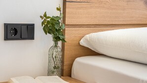 A neatly made bed with a white pillow, a bedside table with an open book, a glass vase with a plant, and a wall socket. - Apartment "Sunnig - Sonennschein" with Mountain View, Balcony & Wi-Fi (Völlan)