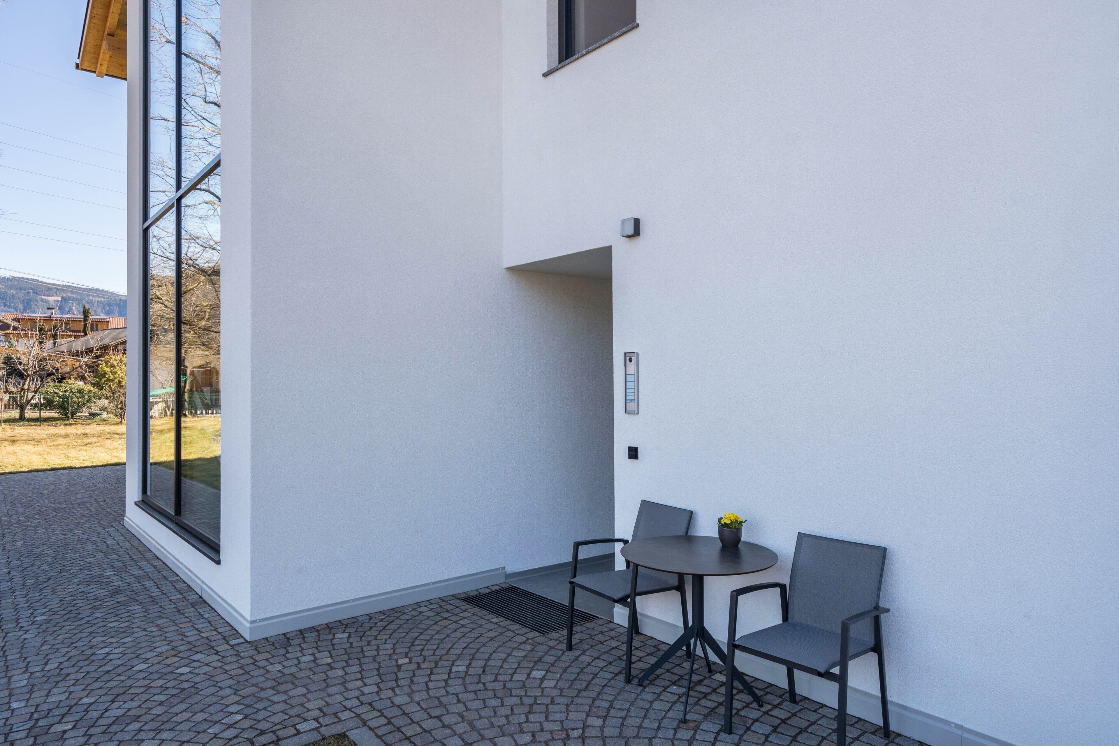 A modern building exterior with a patio area featuring a table and chairs, a large window, and a view of the surrounding landscape.