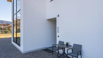 A modern building exterior with a patio area featuring a table and chairs, a large window, and a view of the surrounding landscape.