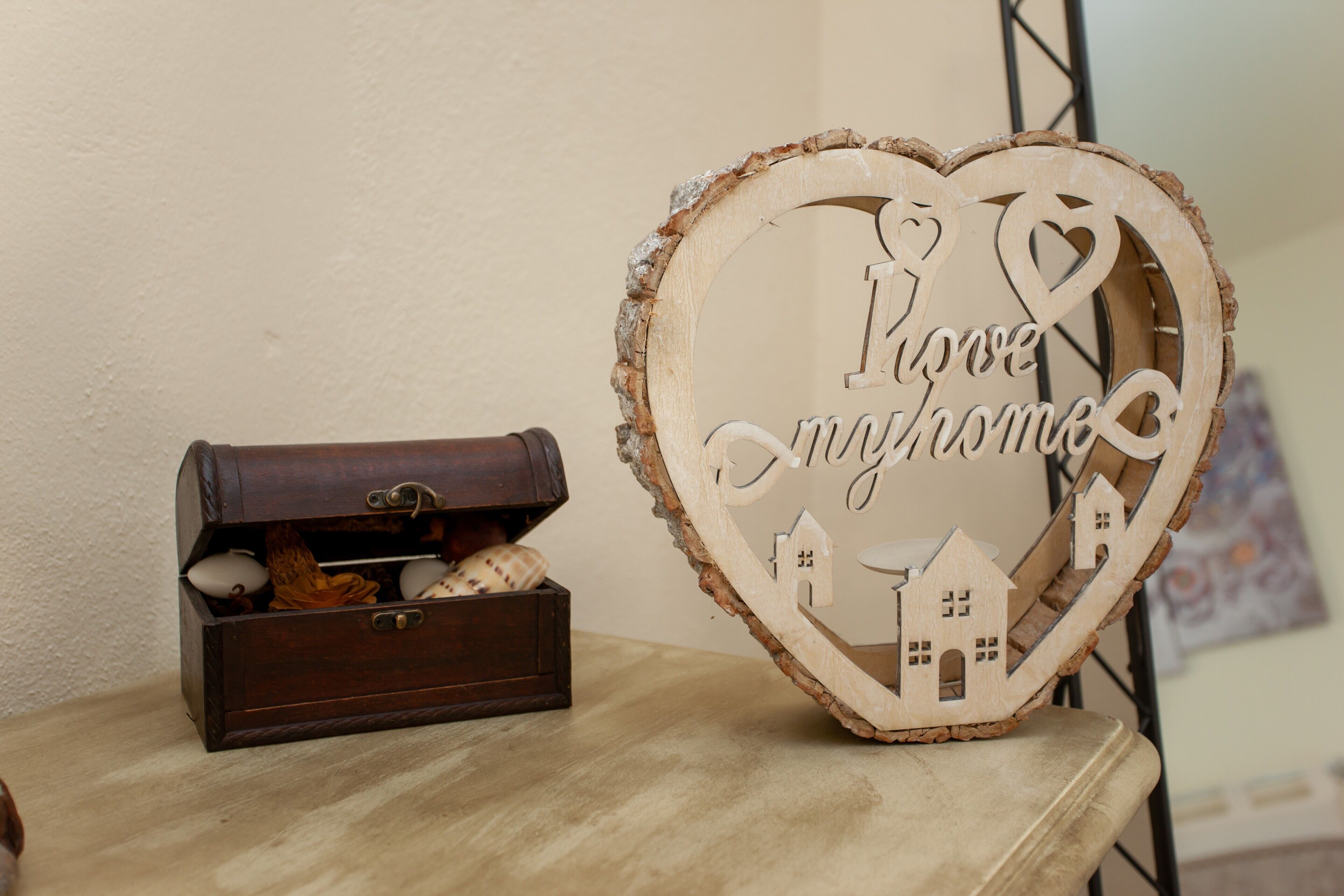 A wooden heart-shaped sign with the words 'Love Home' and small house cutouts, placed on a table next to a dark wooden chest with decorative items.