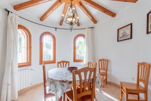A dining area with a round table, six wooden chairs, a chandelier, and three arched windows with curtains. - Villa 'Merced Planta Baja' with Mountain View, Private Pool and Wi-Fi (Calpe)
