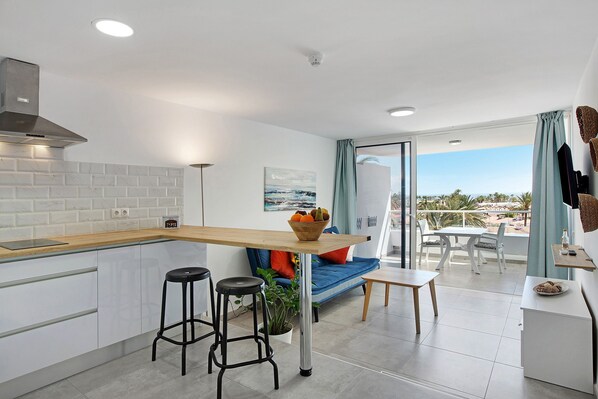 Une cuisine moderne avec un plan de travail en bois, deux tabourets de bar, un coin repas avec une table et des chaises, et un balcon avec vue.