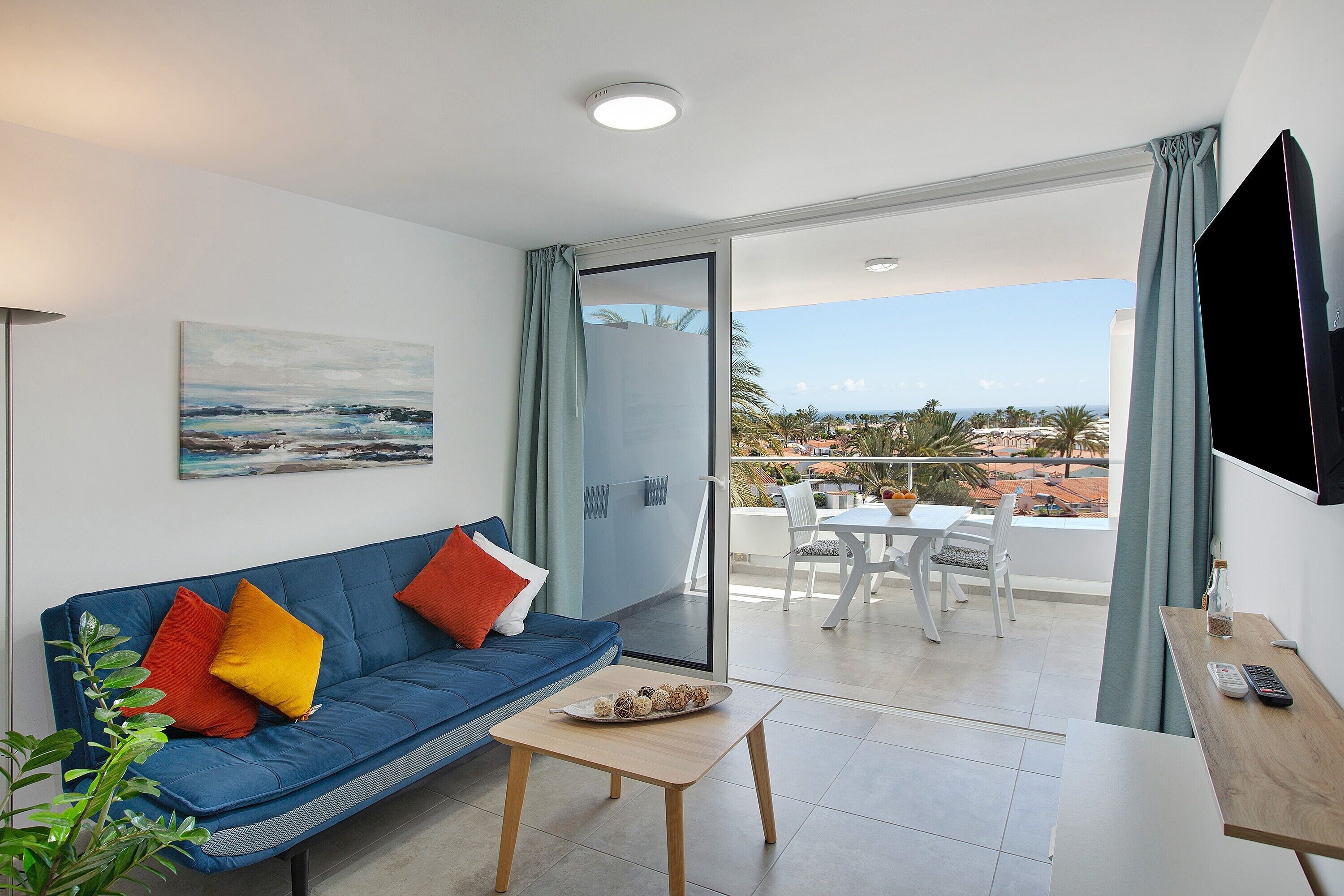 Un salón moderno con un sofá azul, una mesita, un televisor y un balcón con vistas.