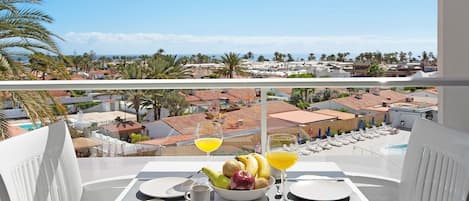 A balcony with a table set for breakfast, including fruits and drinks, overlooking a residential area with palm trees and houses.