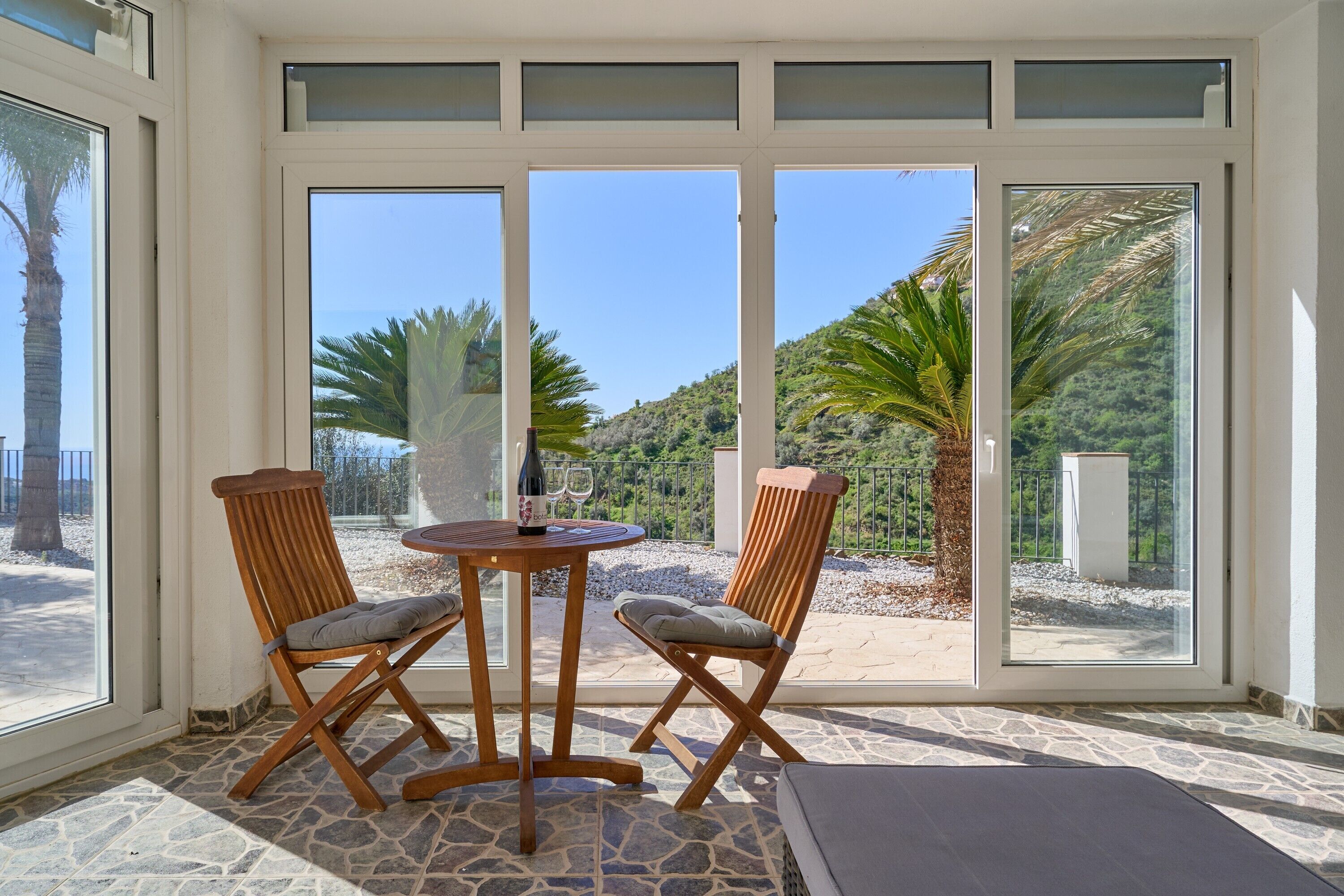 Quarto com piso de pedra, duas cadeiras de madeira, uma mesa redonda e grandes portas de vidro que dão para área externa com palmeiras e vista de uma colina.