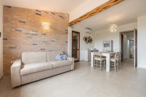 A living room with a brick wall, a sofa, a dining table with chairs, and a wooden ceiling beam.