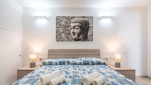 Une chambre à coucher avec un lit, deux lampes de chevet, un tableau représentant un Bouddha accroché au mur, et un lit parfaitement fait avec des serviettes pliées.