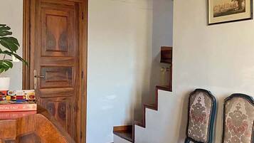 Una puerta de madera, una escalera y dos sillones tapizados en una habitación con paredes blancas y suelo de baldosas.