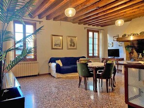 Ein gemütliches Wohnzimmer mit einem blauen Sofa, Holzbalkendecke, einem Esstisch mit Stühlen und einem Fernseher.