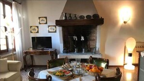 A dining area with a round table set for four, surrounded by green upholstered chairs. There is a fireplace with decorative items above it, and a television on a stand to the left. - Ca' Nadia Apartment  (Venice)