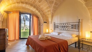 Un dormitorio con techo abovedado de piedra, una cama con cabecero de metal, un armario de madera y vistas al exterior a través de un ventanal.