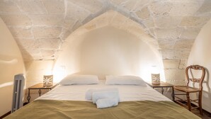 Une pièce avec un plafond en pierre, un lit recouvert de draps blancs, une chaise en bois et une petite table avec une lampe.