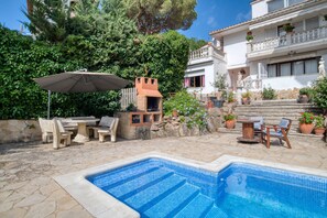 Zona de piscina amb paviment de pedra, barbacoa, mobiliari exterior i un edifici blanc amb balcons.