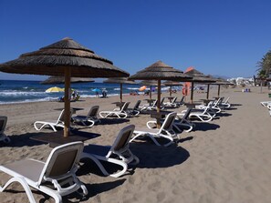 A beach with rows of white lounge chairs and thatched umbrellas, a clear blue sky, and the ocean in the background. - 'Marbella Penthouse apartment' with Sea View, Pool, Wi-Fi, Garden & Terrace (Elviria)