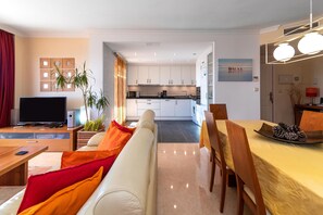 A modern living room with a sofa, TV, and dining area. A kitchen is visible in the background.
