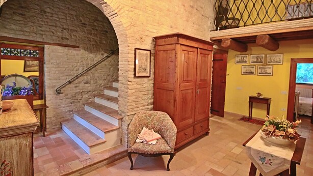 Une pièce dotée d’une arche en pierre, d’un escalier en bois, d’une armoire en bois, d’un fauteuil à motifs floraux et d’une table sur laquelle est posé un vase.