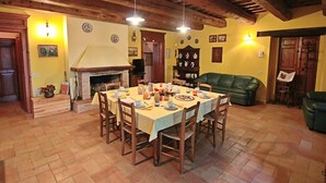 On aperçoit un coin repas avec une table en bois dressée pour le repas, entourée de chaises. Une cheminée est visible au fond, et un canapé vert est placé contre le mur.