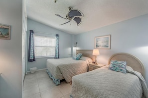 A bedroom with two beds, a ceiling fan, a window with curtains, a bedside lamp, and a wall-mounted artwork.