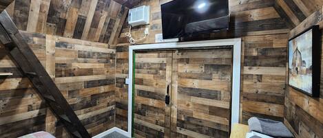 Una habitación con paredes de madera, un televisor de pantalla plana sobre una puerta, una escalera, una cama con cubrecama estampado y una mesita con toallas.