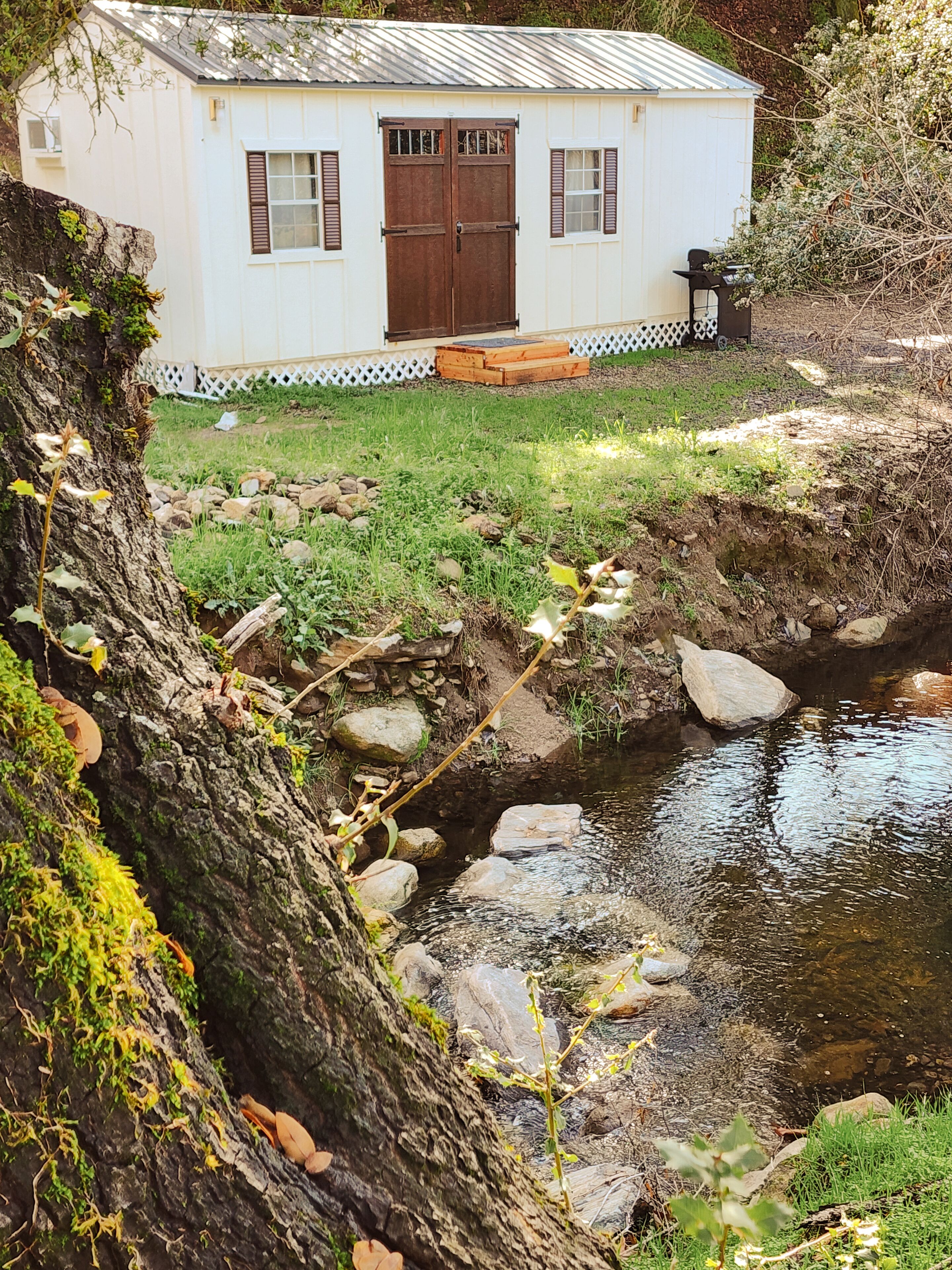 Una pequeña cabaña blanca con puerta y ventanas marrones, rodeada de césped y un arroyo.