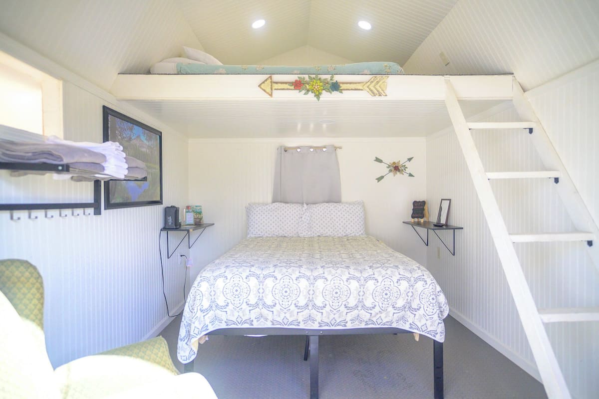 A bedroom with a bunk bed, a bed with a patterned bedspread, a ladder, a shelf with a picture frame, and a small table with a lamp.