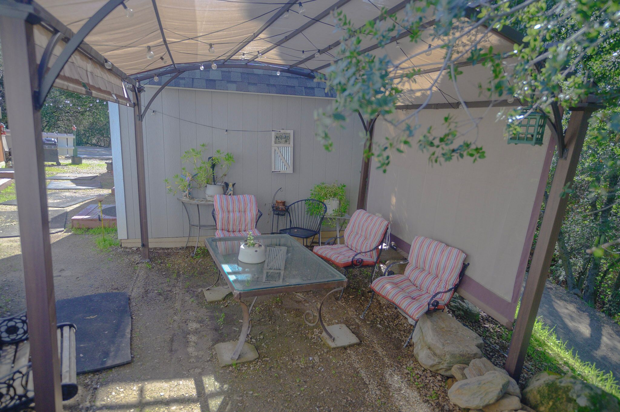 Ein überdachter Patio-Bereich mit einem Glastisch und gestreiften Stühlen, umgeben von Topfpflanzen und einem kleinen Schuppen.