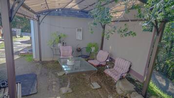 Ein überdachter Patio-Bereich mit einem Glastisch und gestreiften Stühlen, umgeben von Topfpflanzen und einem kleinen Schuppen.