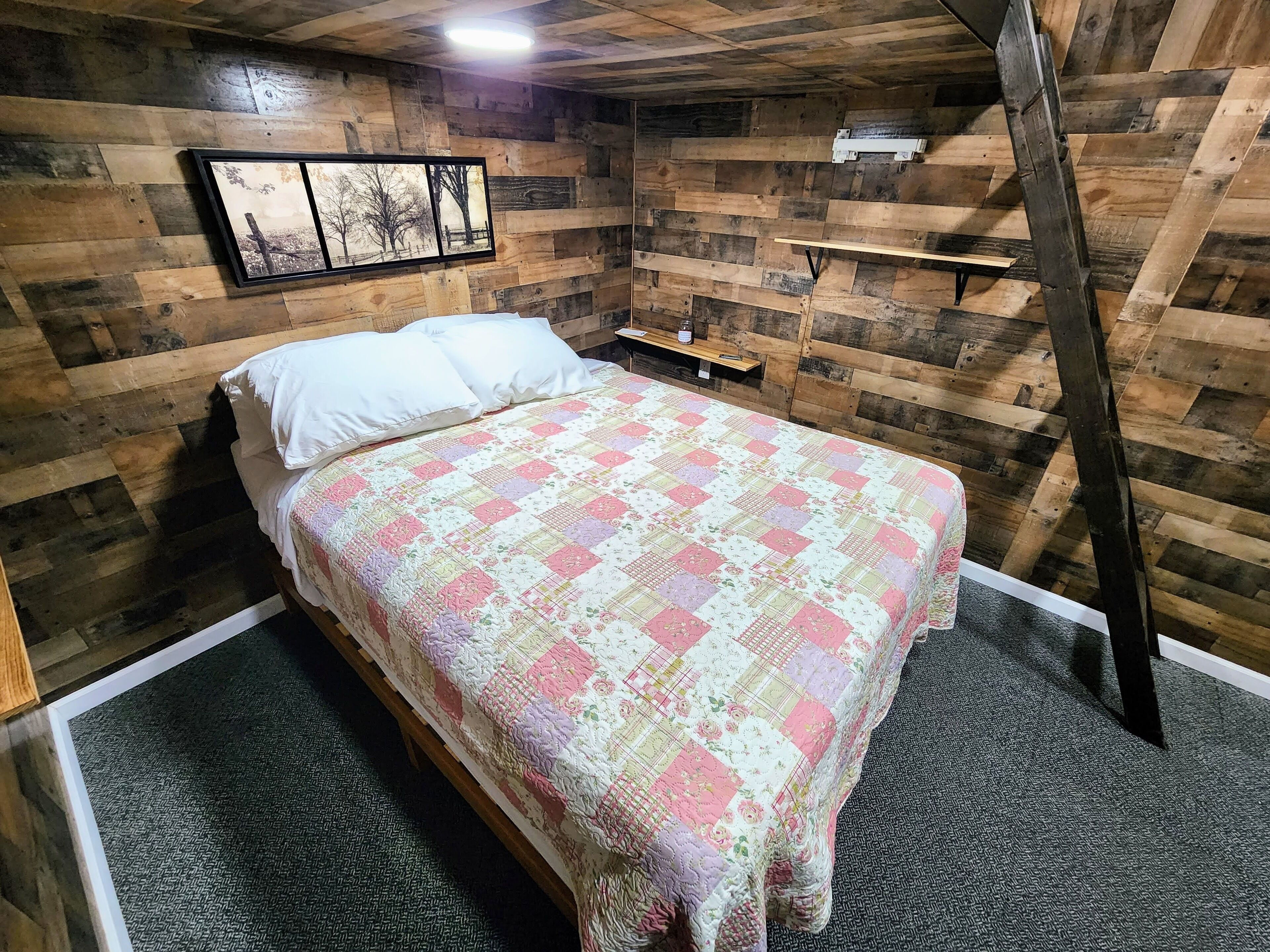 A cozy bedroom with a wooden sloped ceiling, a bed with a floral quilt, a nightstand, and a wall-mounted shelf.
