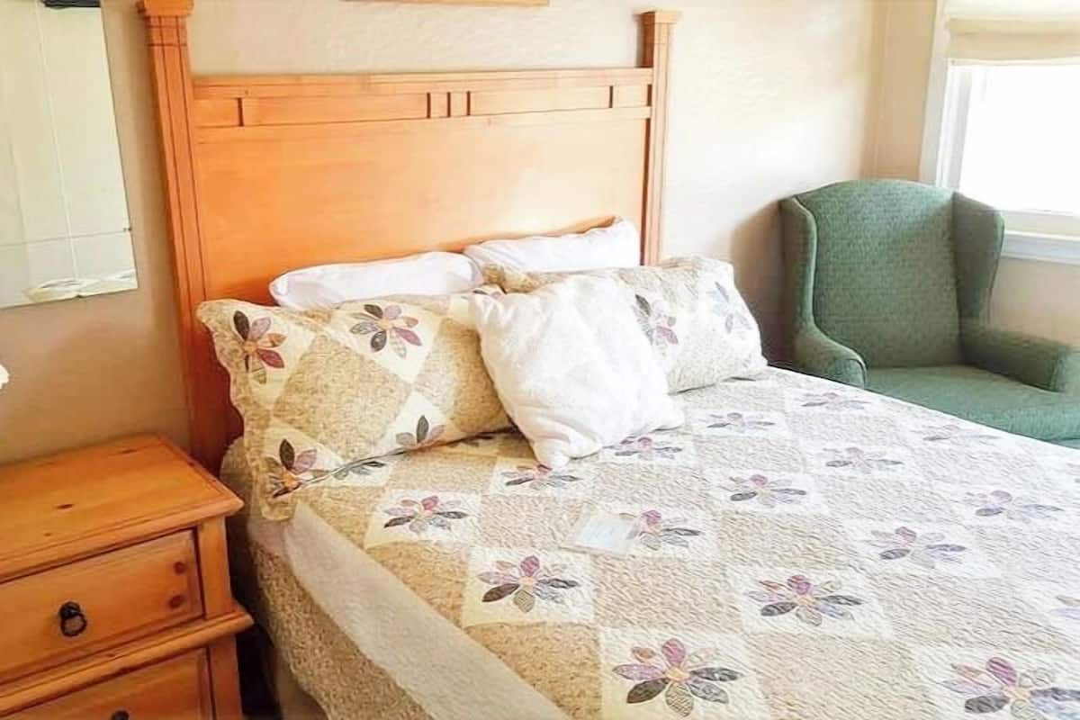 A bed with a floral quilt, a wooden nightstand with a lamp, and a green armchair.