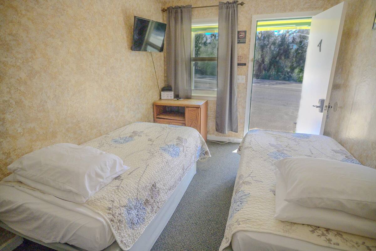 A room with two beds, a small wooden table, a television on the wall, and a door leading outside.