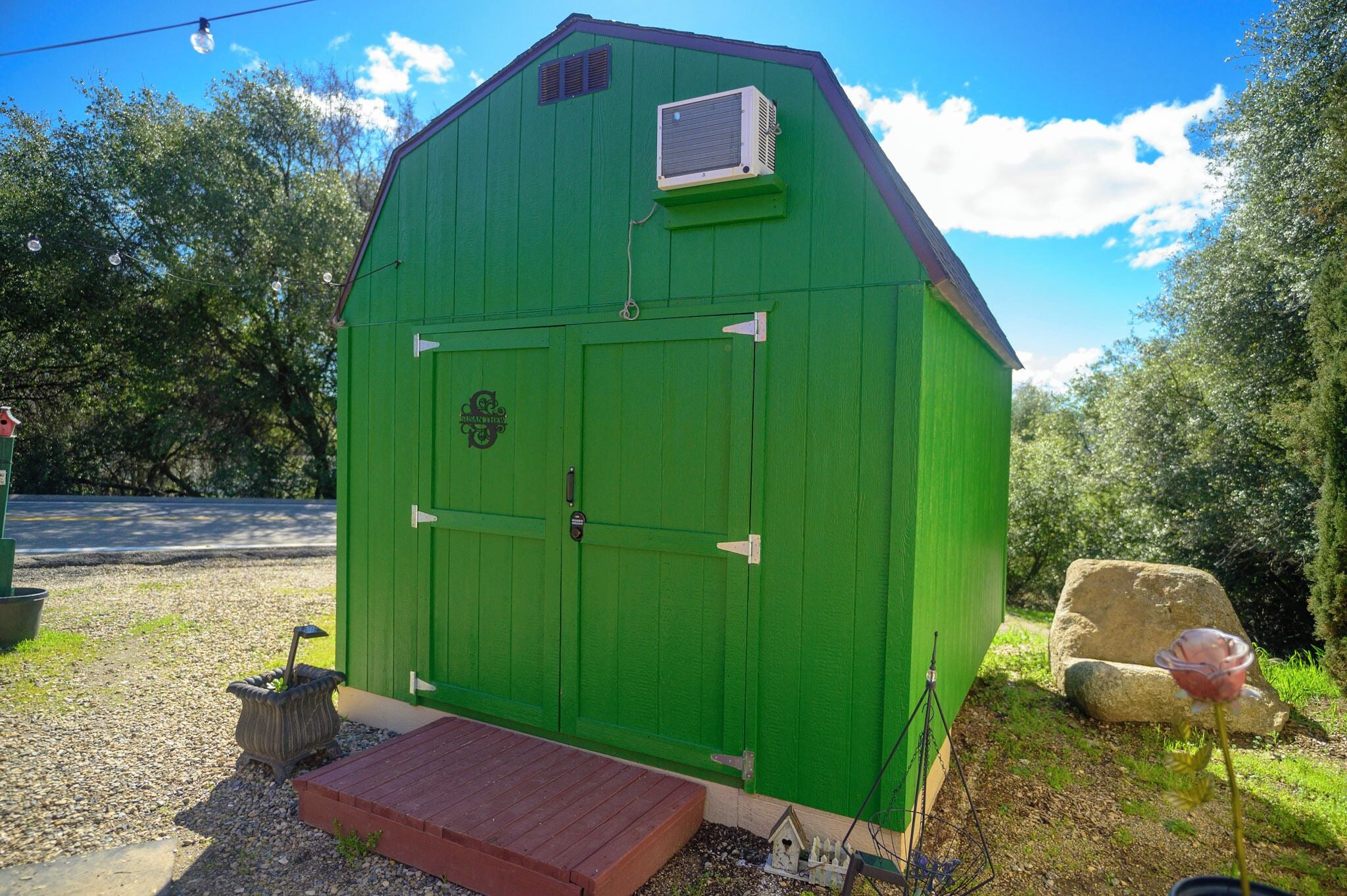 A green shed with a small porch, an air conditioning unit, and a decorative flower on the right.